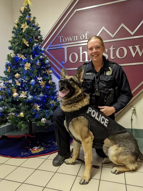 Office Kehr and her K9 Vasco, kneel in front of a christmas tree an the Town of Johnstown logo for a photo highlighting the K9s new police vest.